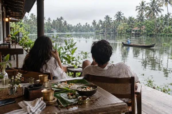 Two travellers linger over a banana leaf breakfast on a verandah above the Kumarakom backwaters as a canoe glides past