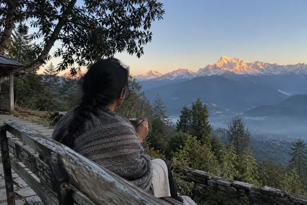 A woman in a wool shawl holds a cup of chai on a bench, facing the Panchachuli peaks at dawn in Munsiyari